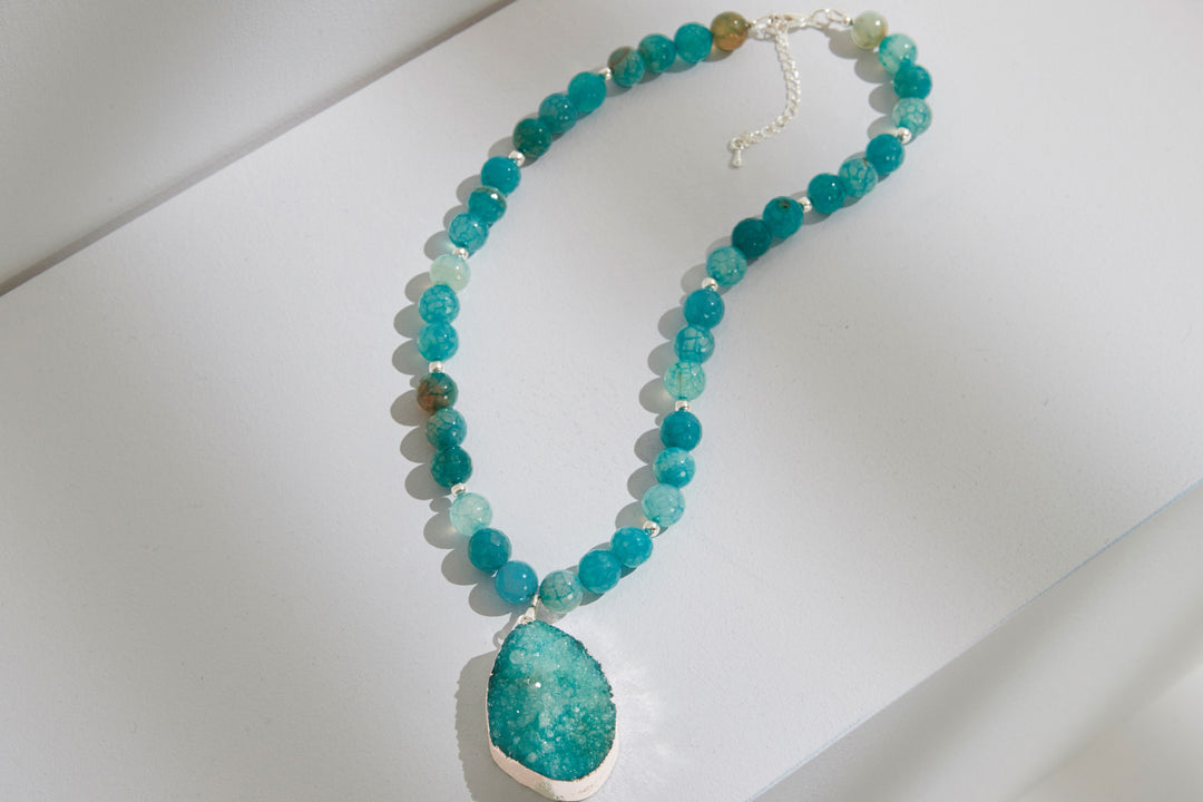 Lily Ella Collection turquoise beaded necklace with silver clasp and textured pendant displayed on white surface, elegant handcrafted jewelry accessory.