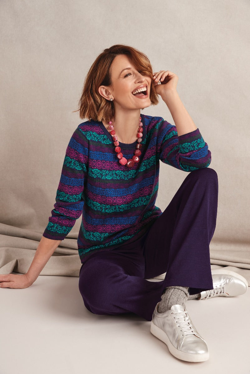 Lily Ella Collection stylish woman wearing vibrant purple and blue patterned sweater with matching purple trousers, fashionable silver sneakers, and pink beaded necklace, smiling in a casual seated pose.