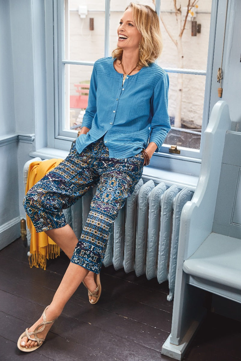 Lily Ella Collection stylish woman wearing blue button-up shirt and patterned trousers with gold sandals sitting by window with blue interior decor