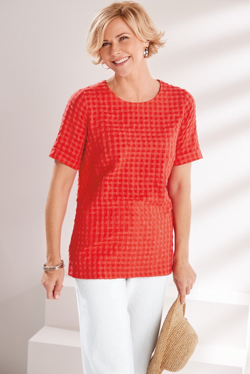 Lily Ella Collection red checkered pattern tunic top, stylish mature woman in a casual summer outfit with white pants and a straw bag.