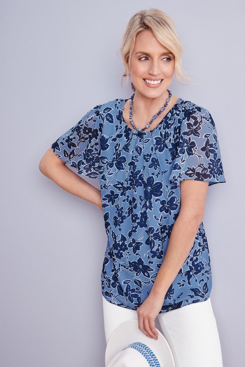Lily Ella Collection blue floral blouse for women, stylish short-sleeve top with navy and white flower print, paired with white trousers and casual accessories, modern outfit for spring and summer.