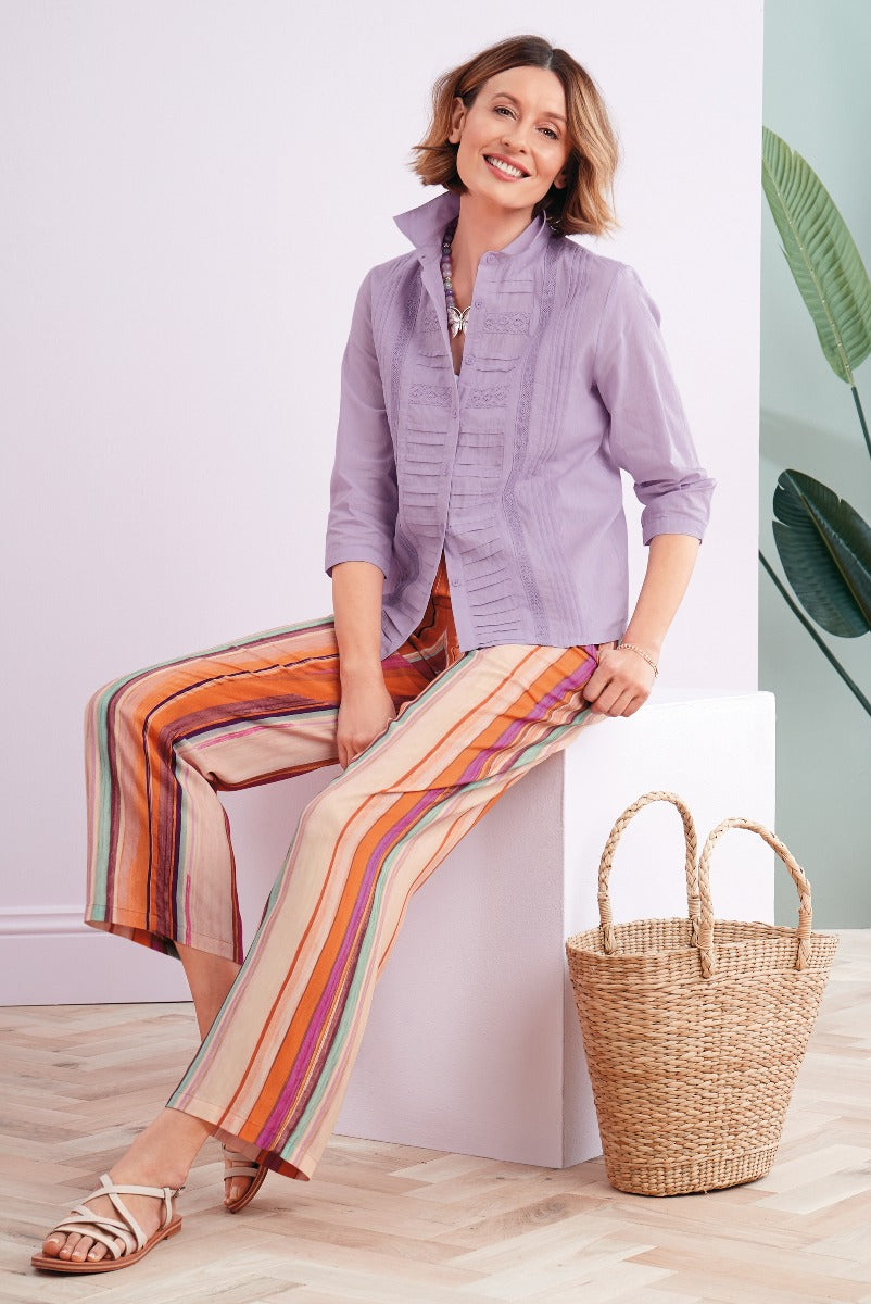Lily Ella Collection stylish lavender blouse with detailing and multicolor striped palazzo pants, accessorized with tan strappy sandals and a woven basket bag, in a modern indoor setting for a spring/summer chic look