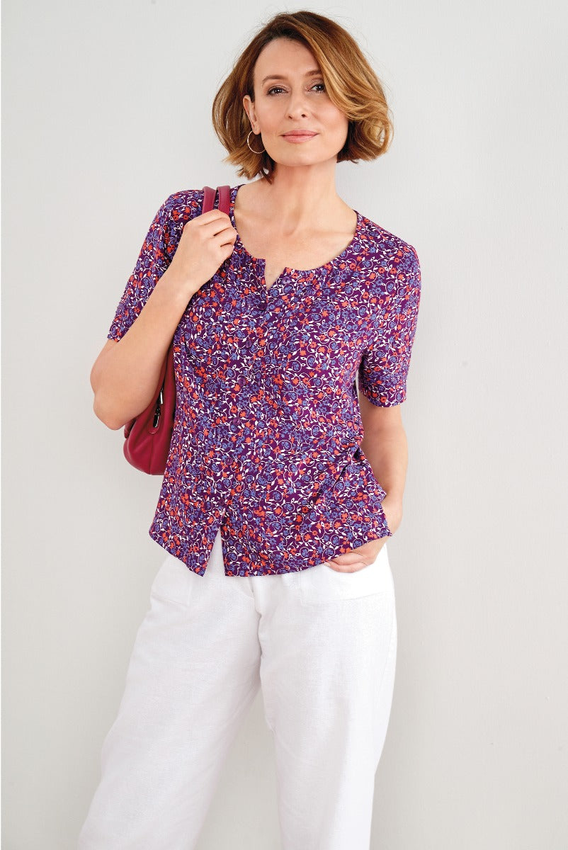 Lily Ella Collection floral print blouse in purple and red tones paired with white trousers, stylish mature woman modeling casual elegant outfit