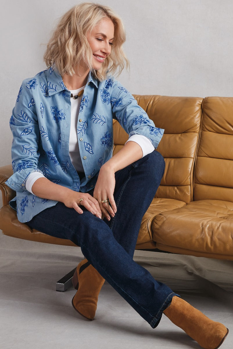 Lily Ella Collection stylish blue floral print shirt, paired with dark denim jeans and brown suede boots, sitting casually on a tan leather couch.