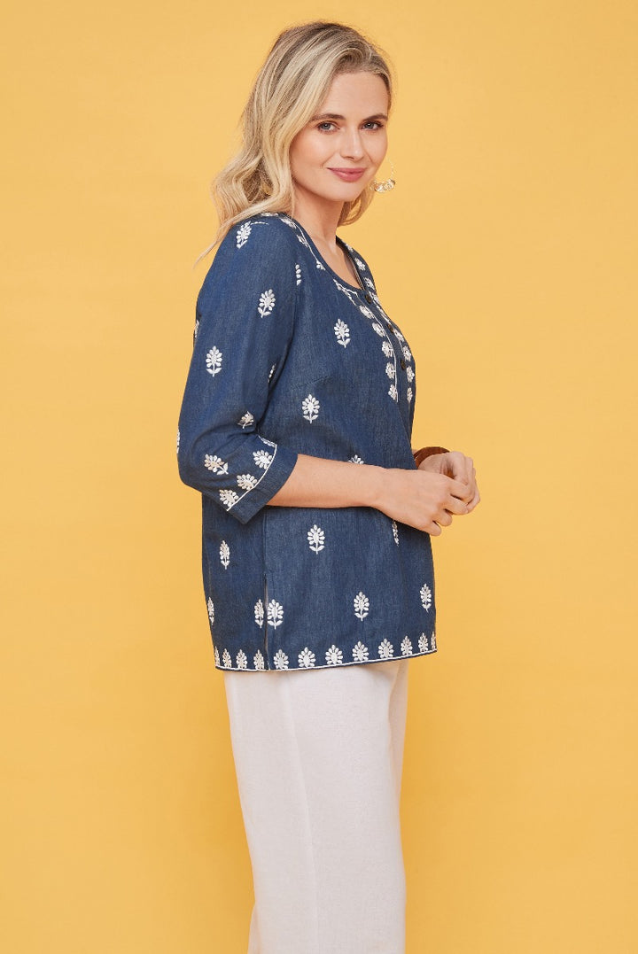 Lily Ella Collection navy blue floral embroidered tunic top with three-quarter sleeves paired with white trousers on model against yellow background.
