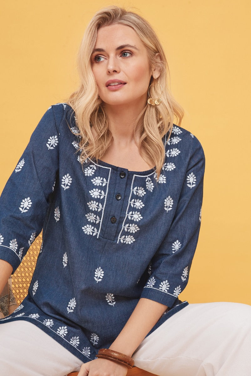 Lily Ella Collection navy blue bohemian tunic with white floral embroidery, three-quarter sleeves, relaxed fit, styled with white pants and gold earrings, warm yellow background.
