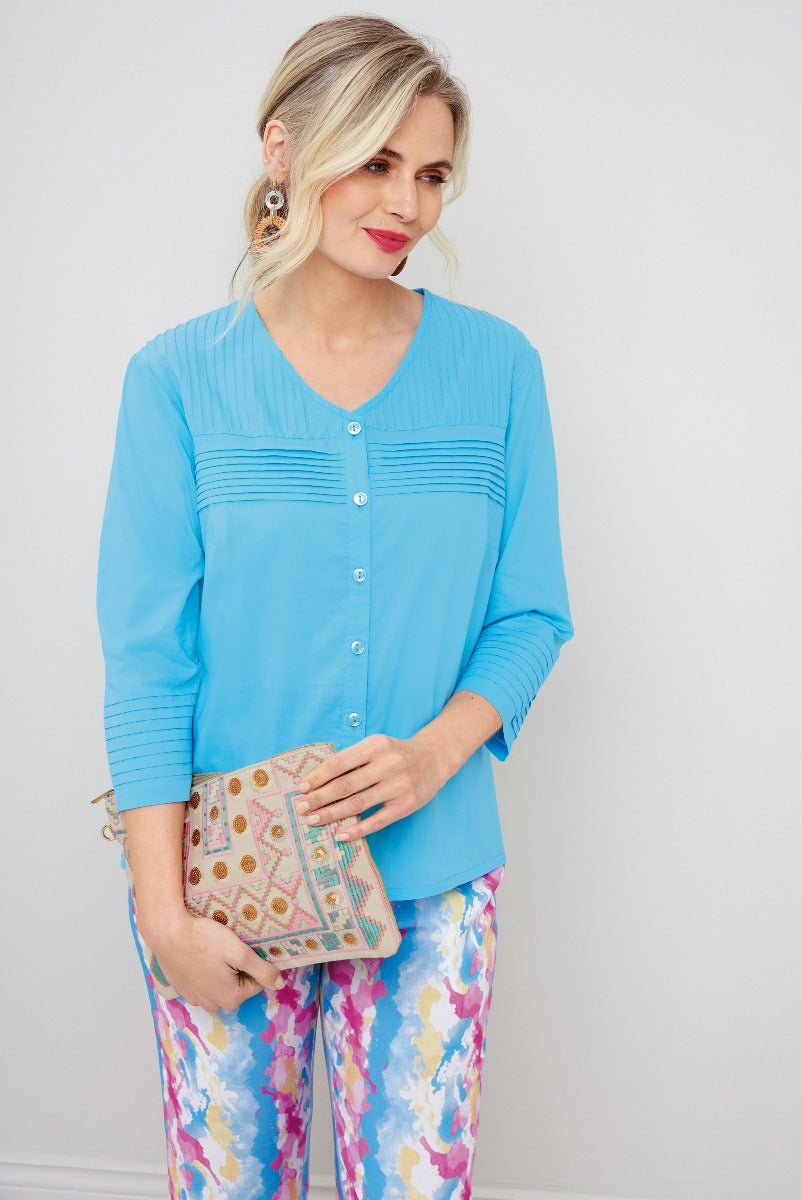 Lily Ella Collection stylish sky blue pleated blouse with button-up front paired with vibrant patterned trousers and accessorized with a pastel embroidered clutch for a chic women's spring outfit.