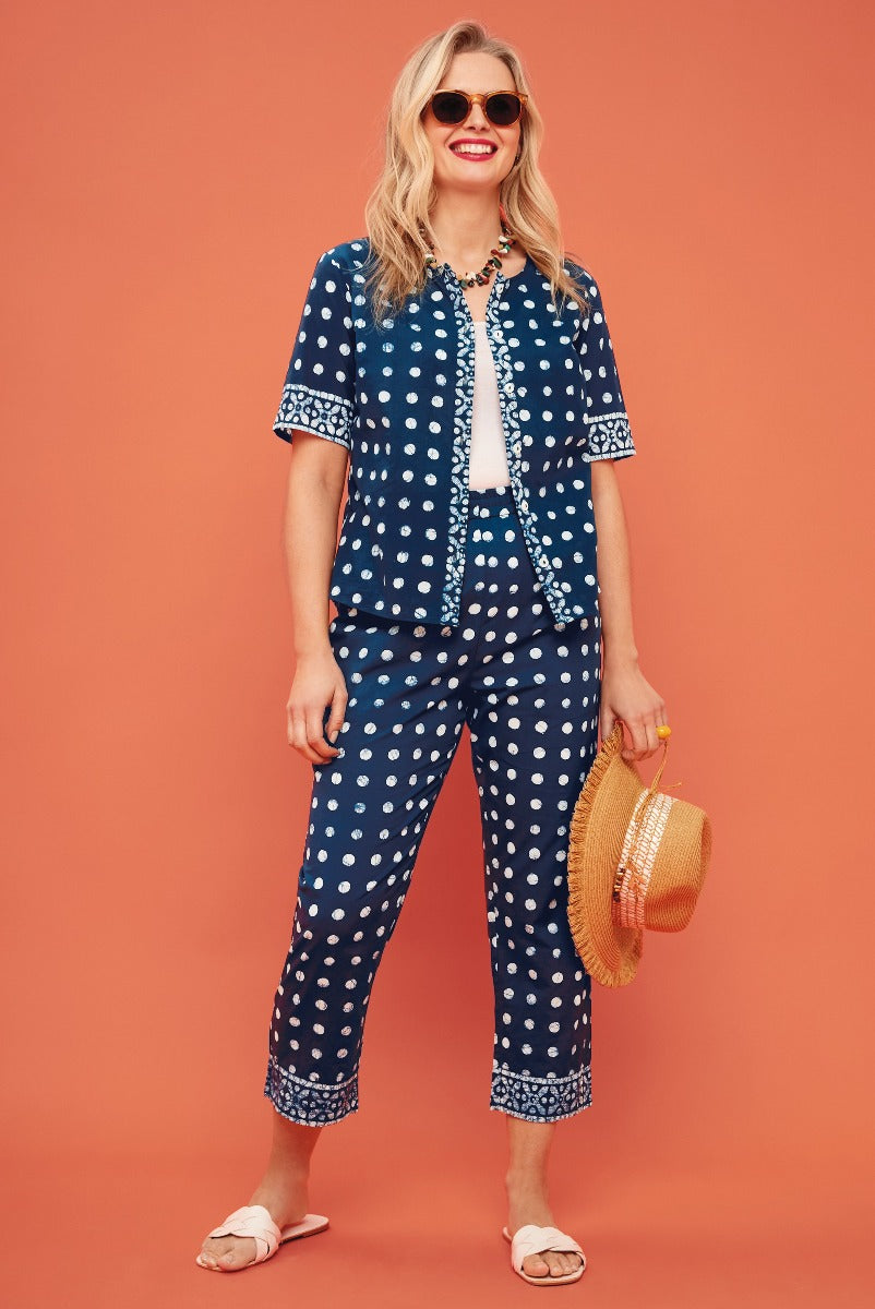 Lily Ella Collection model wearing a navy blue polka-dot patterned pyjama set with a white camisole, accessorized with round sunglasses, a statement necklace, and carrying a straw bag, standing against an orange background.