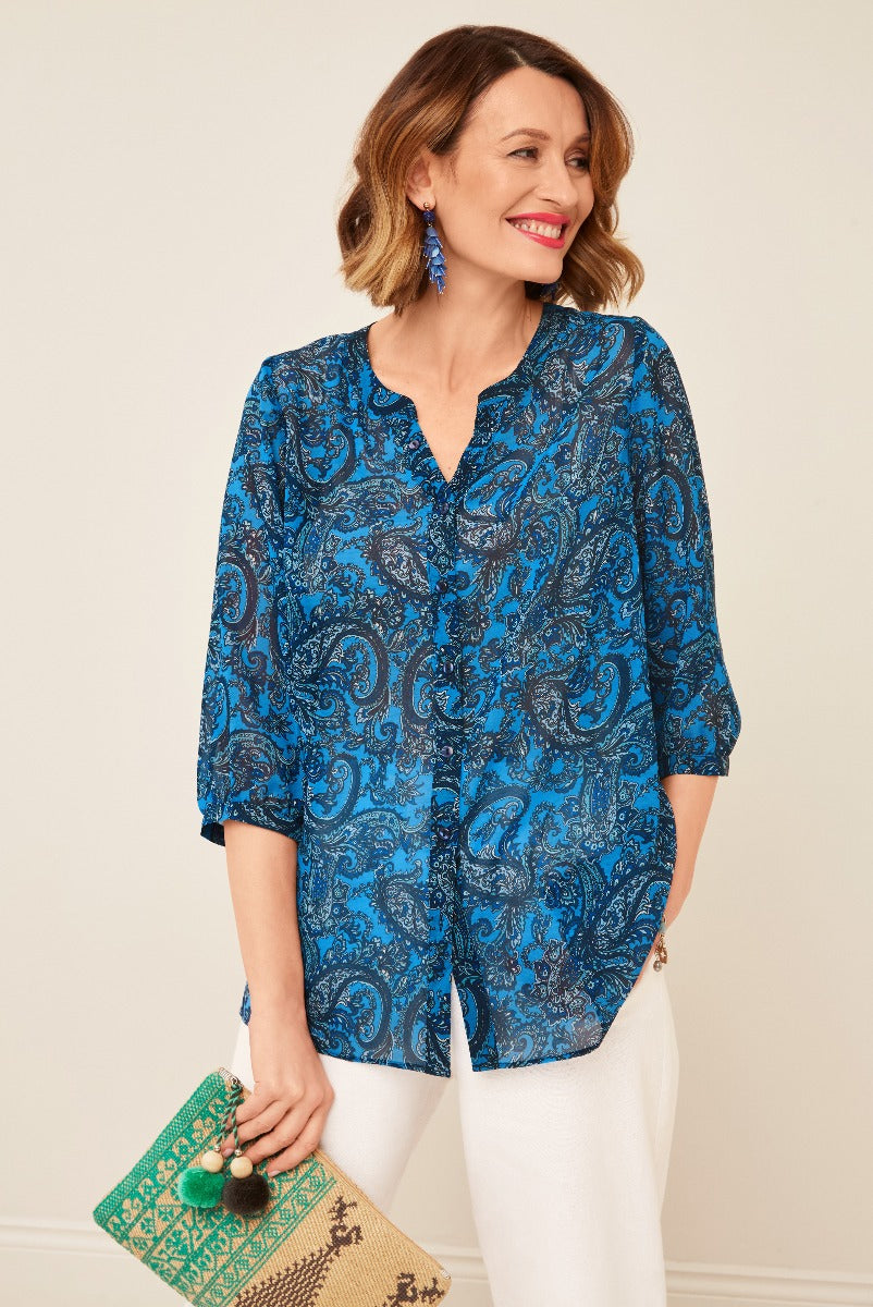 Lily Ella Collection blue paisley print blouse, stylish women's casual top with three-quarter sleeves, paired with white trousers and accessorized with tassel earrings and a patterned clutch bag