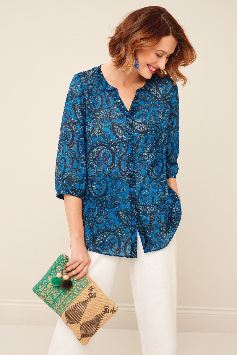 Lily Ella Collection blue paisley print shirt with casual style paired with white trousers and matching blue earrings holding a beige clutch with tropical design for a vibrant summer outfit.