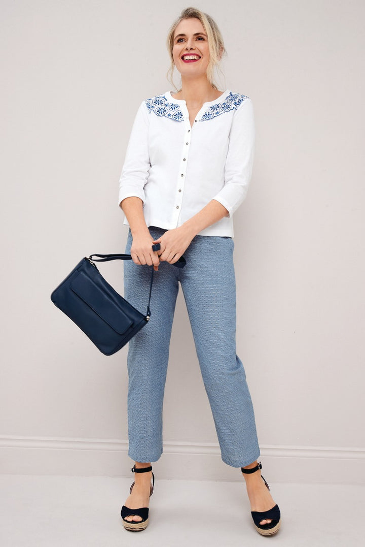 Lily Ella Collection model smiling wearing a blue embroidered white blouse, textured blue trousers, with navy blue shoulder bag and black wedged sandals.