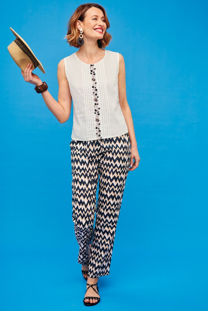 Lily Ella Collection cheerful woman posing in a cream sleeveless top with black floral embroidery and navy and white chevron patterned trousers, holding a straw clutch, against a vibrant blue background