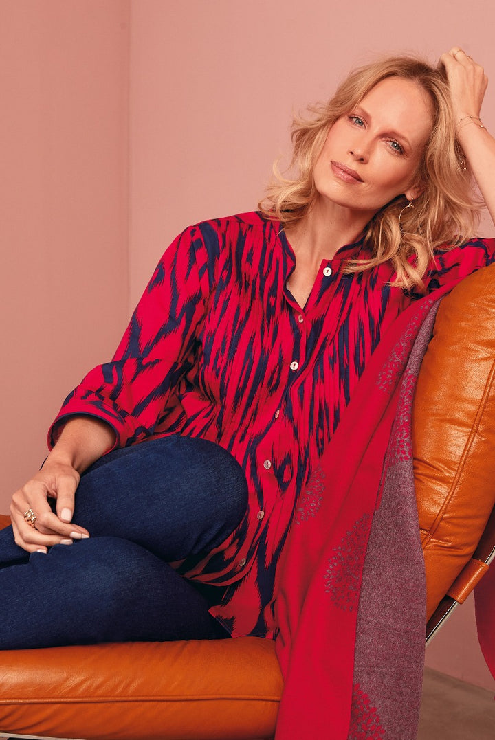 Lily Ella Collection vibrant red and navy abstract print shirt for women, stylish casual wear, paired with denim, model posing with elegance on a tan leather chair
