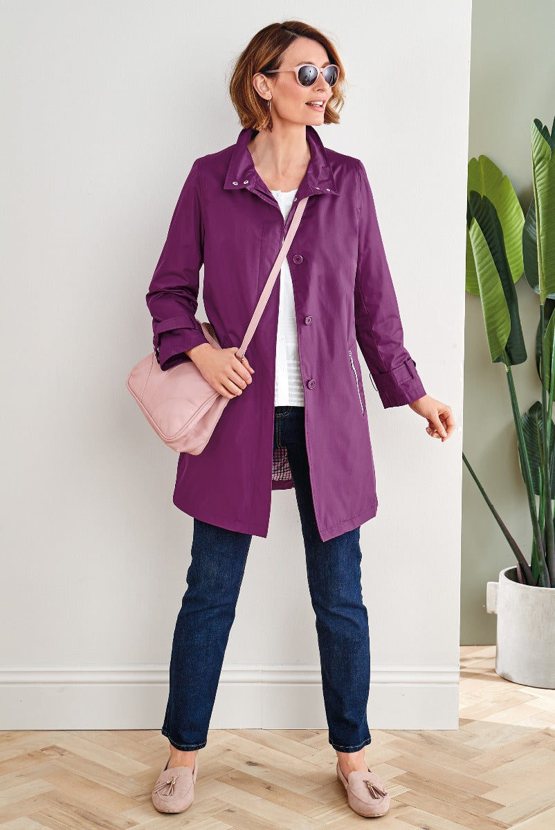 Lily Ella Collection stylish purple raincoat for women, model wearing casual chic outfit with white top and dark denim jeans, accessorized with pastel pink shoulder bag and beige loafers, standing in a modern interior setting.
