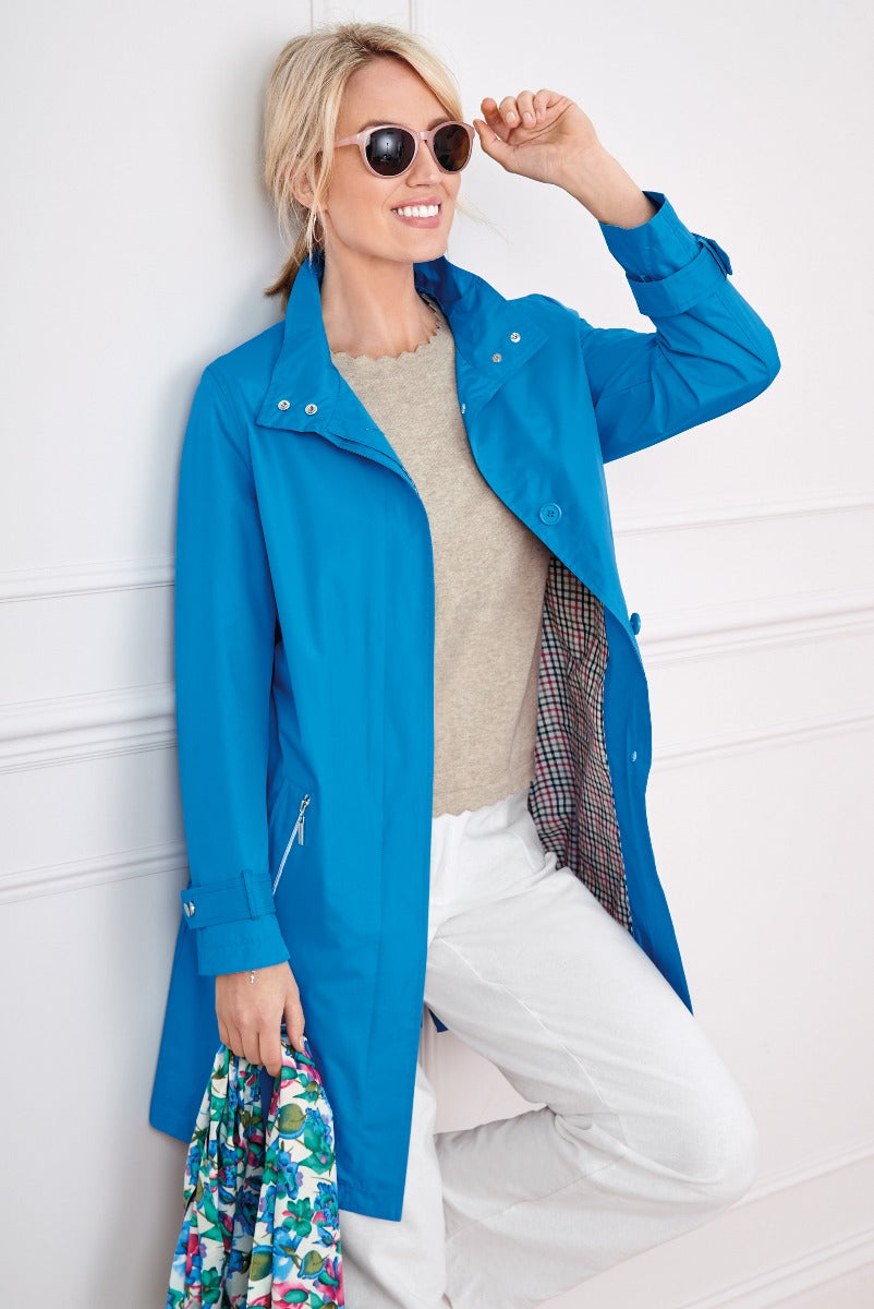 Lily Ella Collection vibrant blue stylish raincoat for women, smiling model wearing casual grey top and white pants accessorized with round sunglasses and holding a colorful floral scarf.