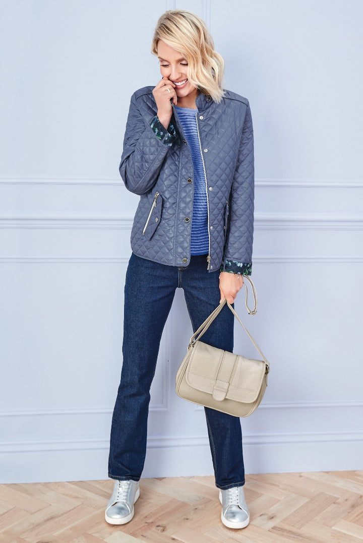 Lily Ella Collection stylish woman smiling wearing blue quilted jacket, patterned blouse, dark denim jeans, beige shoulder bag, and silver sneakers.