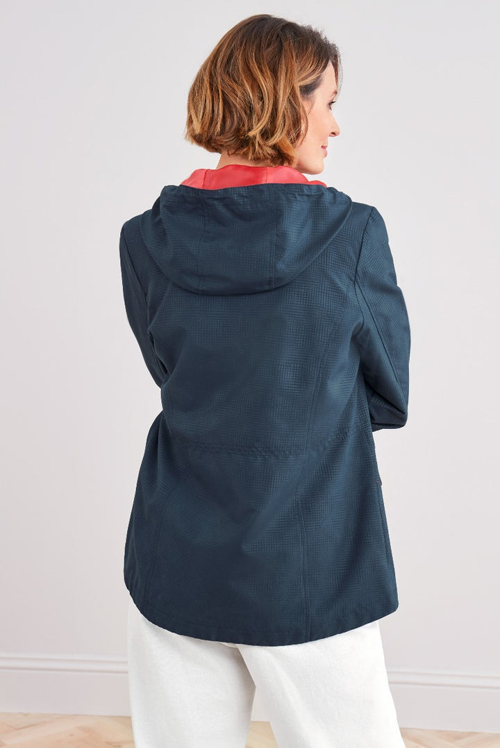 Lily Ella Collection navy blue textured raincoat with coral lining, rear view featuring adjustable hood and hem on model pairing with white trousers