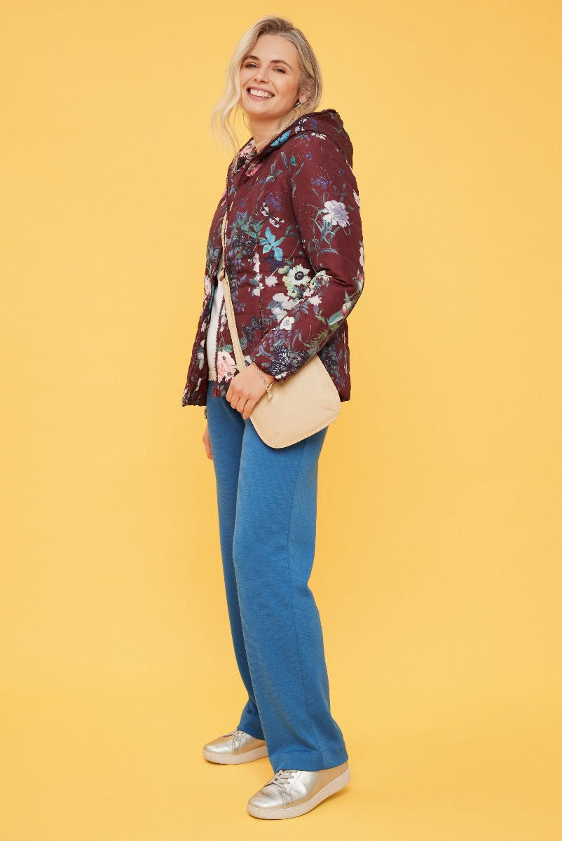 Lily Ella Collection floral patterned quilted jacket in burgundy with casual blue trousers and metallic sneakers on model against yellow background