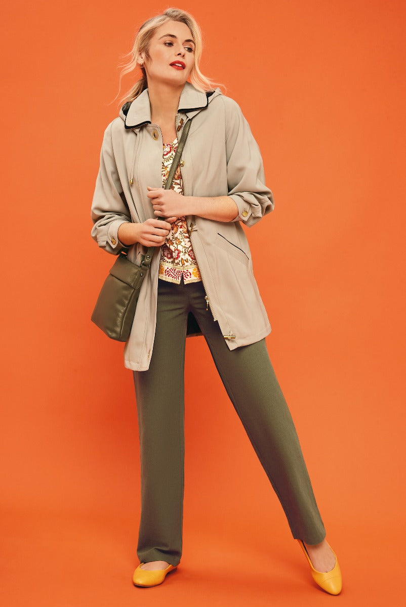 Lily Ella Collection model showcasing a taupe lightweight jacket, patterned blouse, olive green trousers, and mustard yellow flats against an orange background.