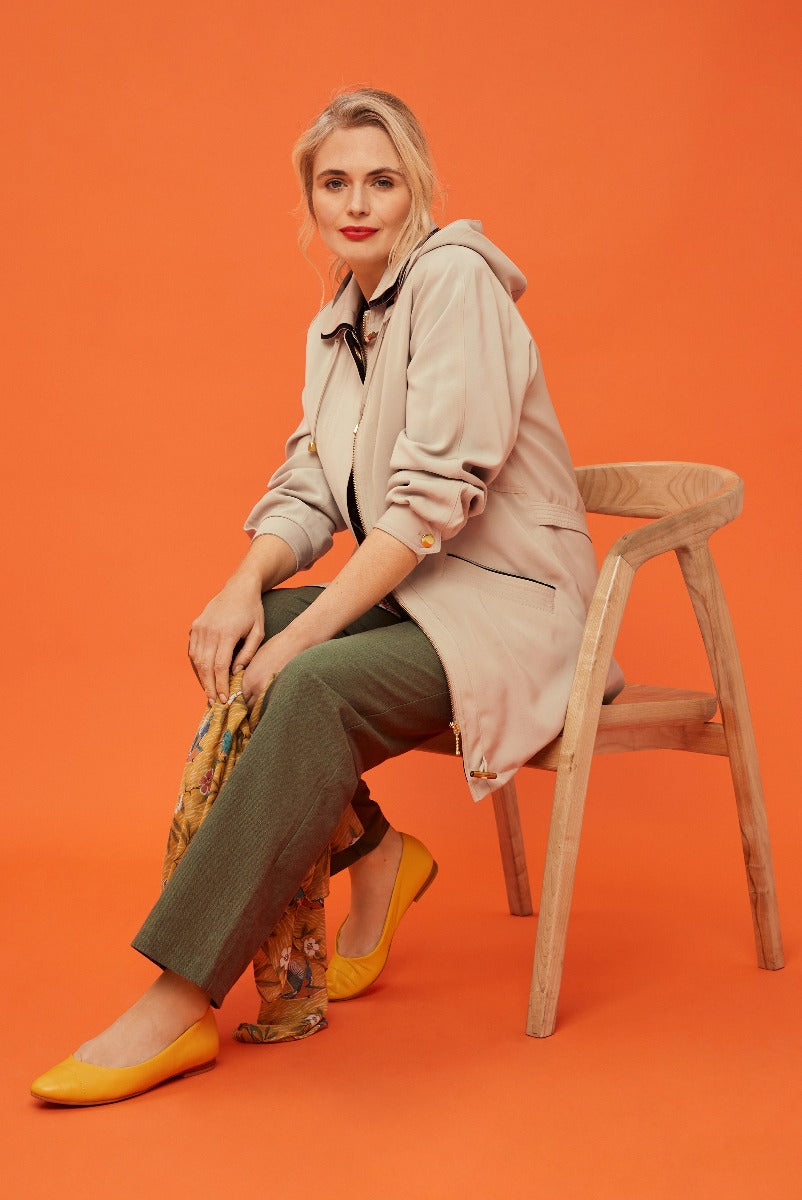 Lily Ella Collection elegant beige trench coat, stylish olive green trousers, floral scarf, yellow flats, woman posing on wooden chair, orange background