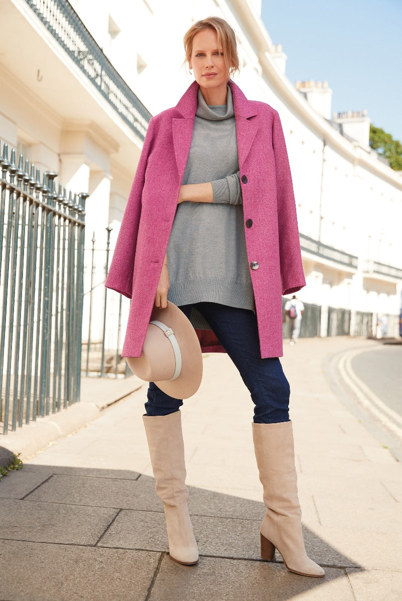 Lily Ella Collection stylish woman in vibrant pink coat, grey turtleneck sweater, dark denim skinny jeans, and beige knee-high boots, holding a light tan hat, urban chic outfit for fall season.