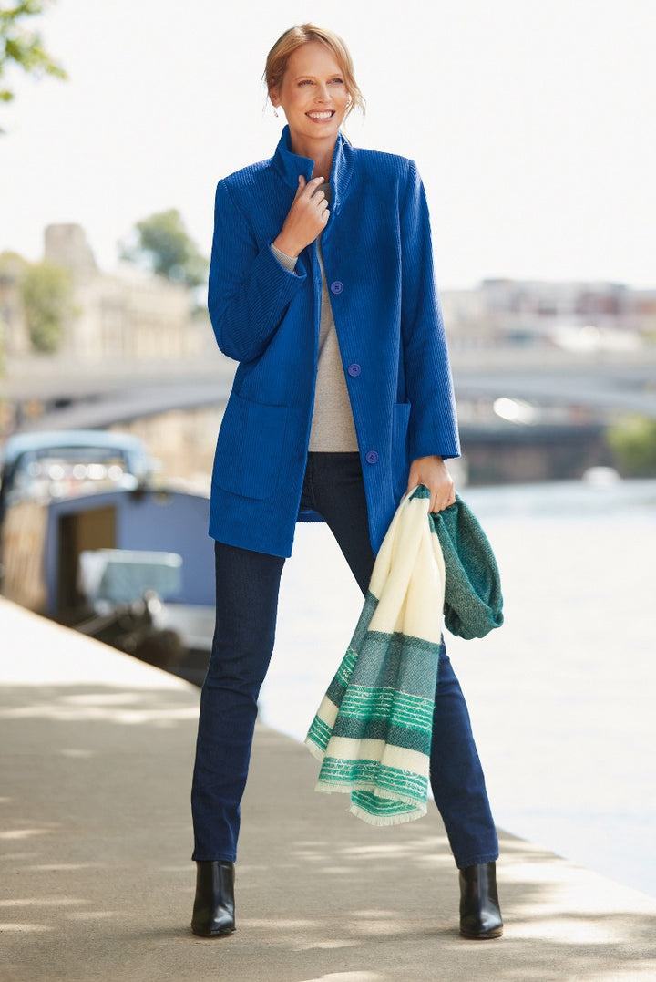 Lily Ella Collection elegant blue buttoned coat for women paired with dark jeans and layered with a grey sweater, model smiling and holding a striped green and white scarf, stylish autumn-winter fashion outdoor look