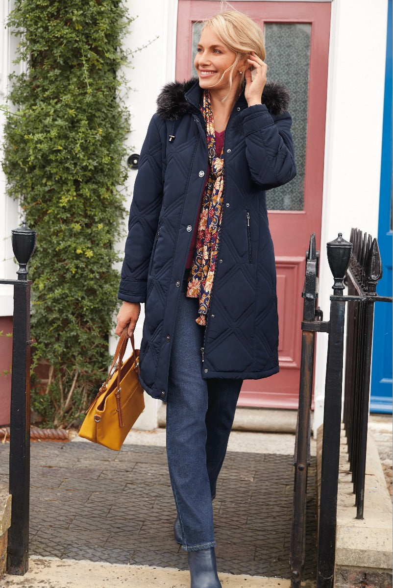 Lily Ella Collection navy blue quilted coat with fur-trim hood, paired with floral blouse, denim jeans, and mustard handbag for a stylish women's autumn outfit.