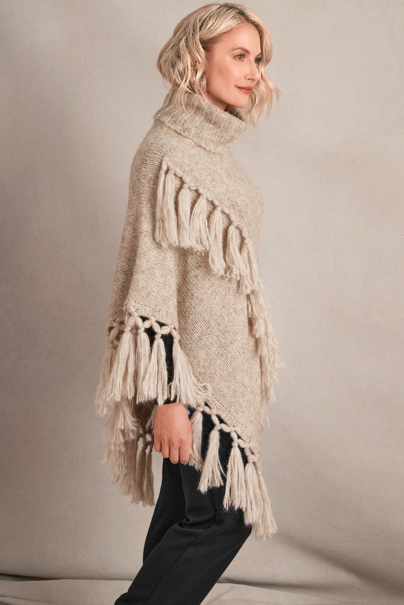 Lily Ella Collection beige tassel poncho with high neck, knit texture, and fringe detailing, styled with black trousers for a chic look