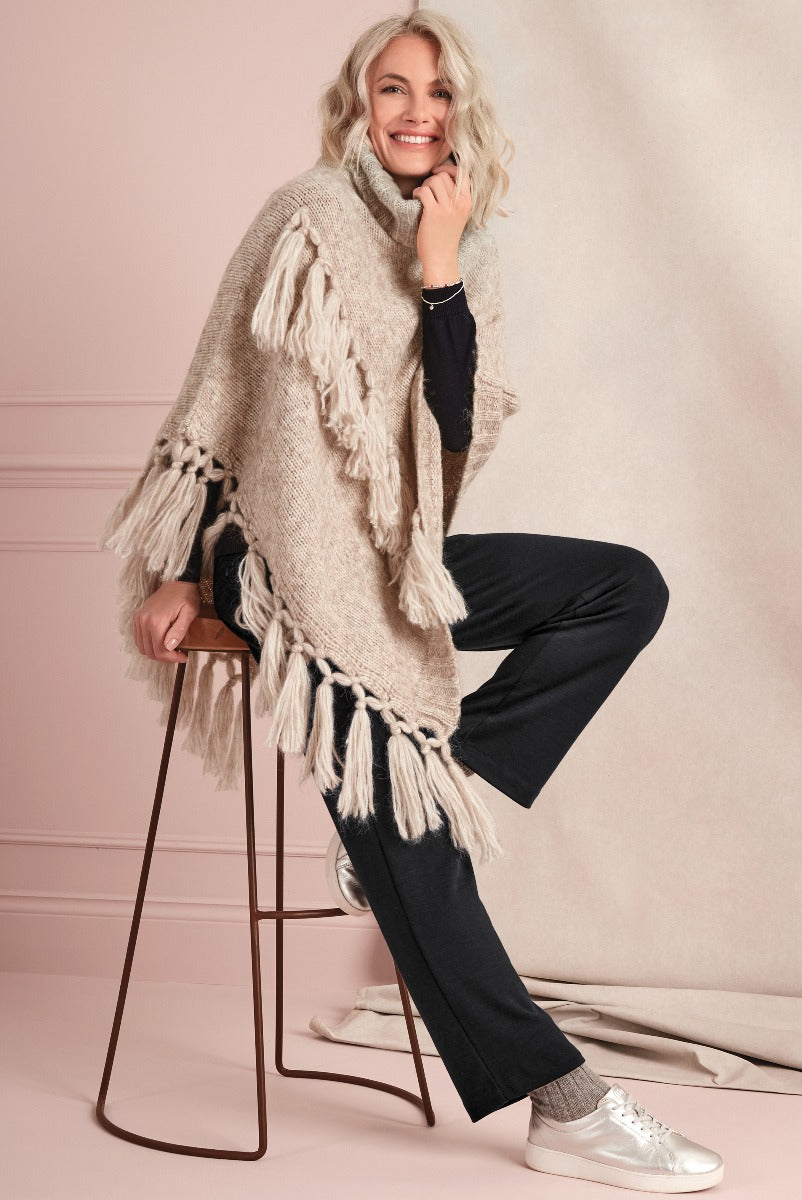 Lily Ella Collection beige fringed poncho with happy smiling woman in casual style, dark trousers and white sneakers against pink backdrop.