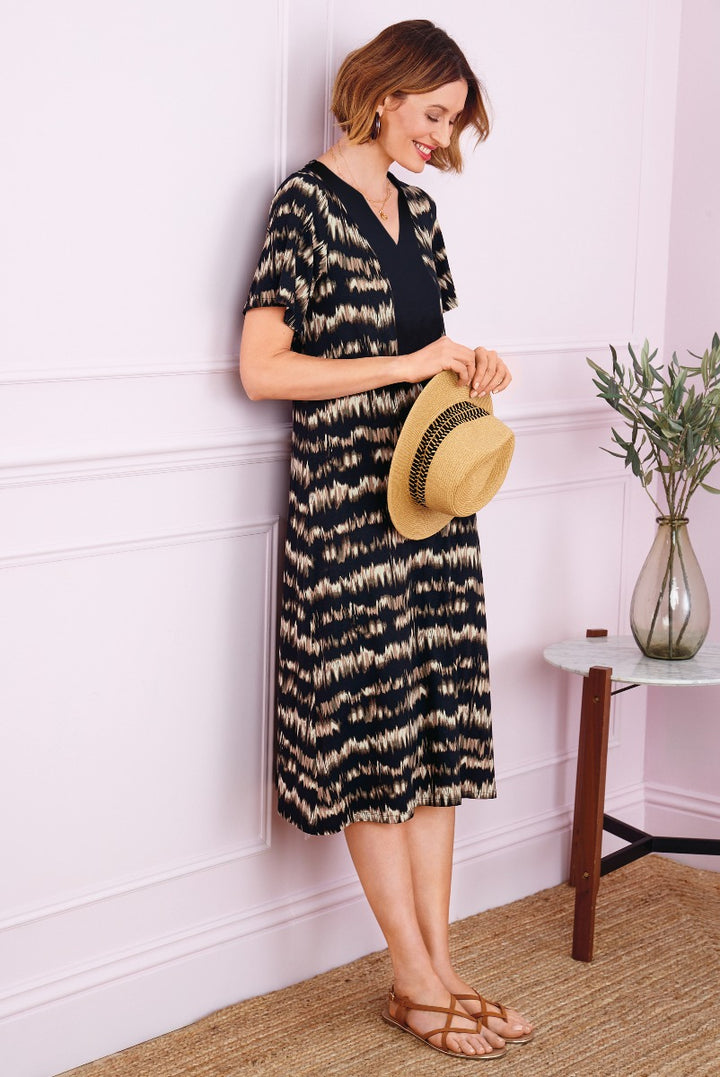 Lily Ella Collection black and tan zigzag patterned dress with short sleeves, V-neck, mid-length style, paired with a straw hat and brown sandals on a smiling model.