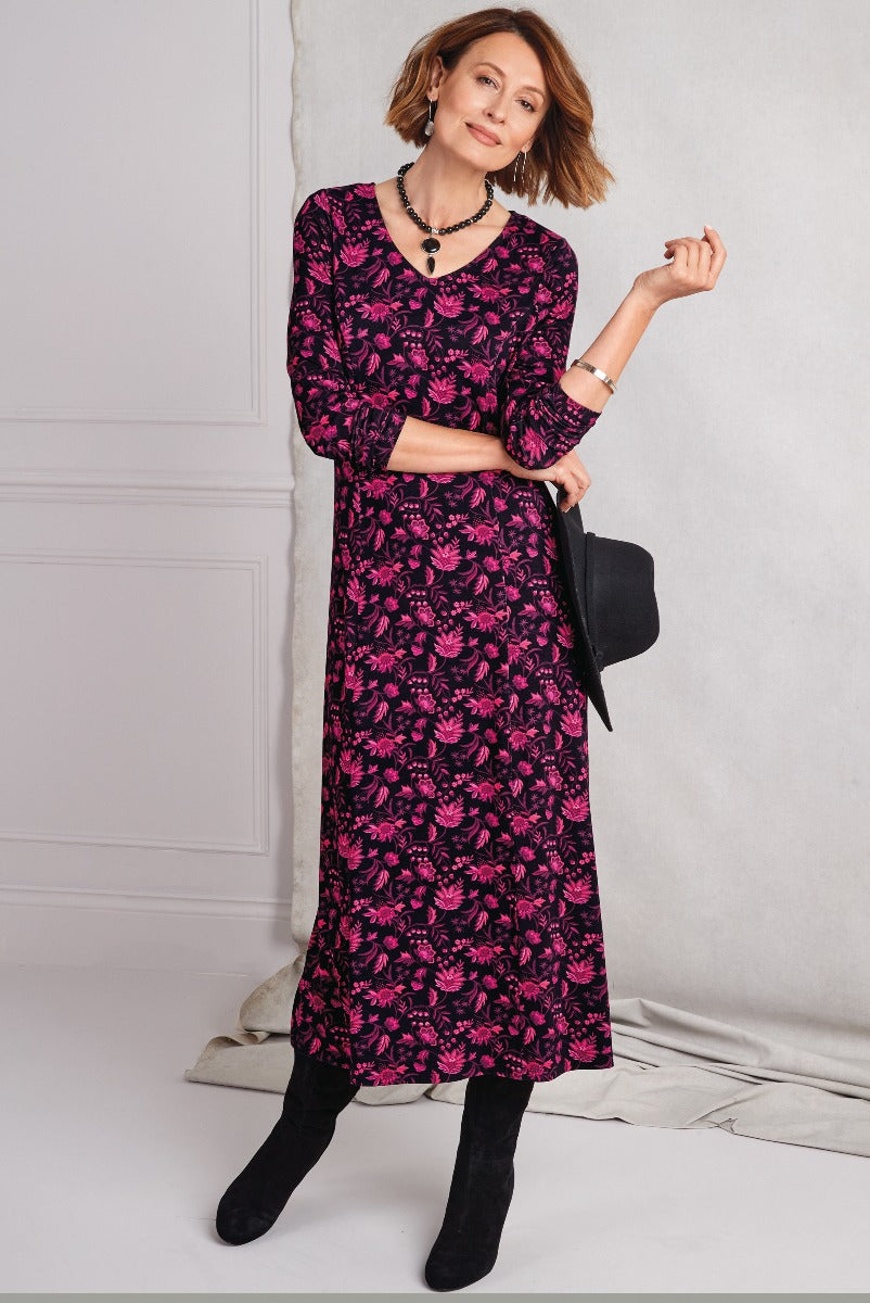 Lily Ella Collection elegant woman posing in a vibrant plum floral midi dress with three-quarter sleeves, perfect for autumn wear, paired with black suede boots and accessorized with a black hat and silver jewelry.