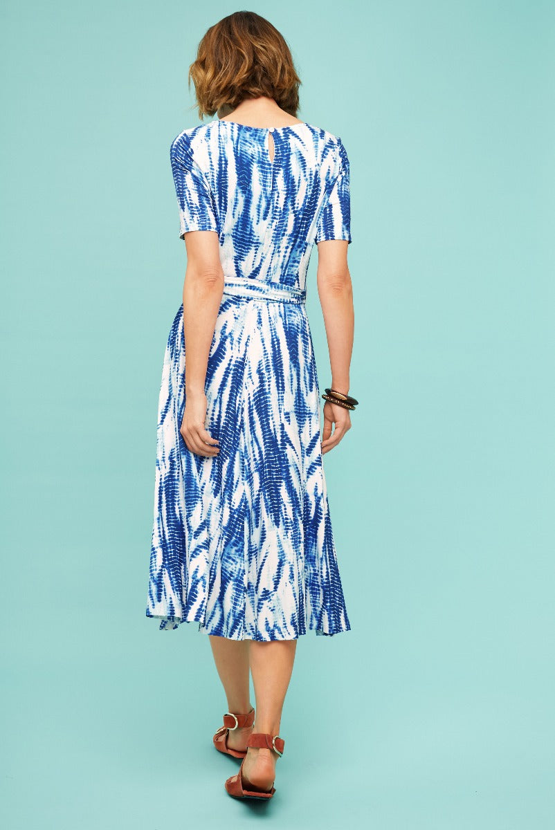 Lily Ella Collection blue and white patterned midi dress with short sleeves and waist tie detail, model poses showing back view, paired with brown strappy sandals on a teal background.