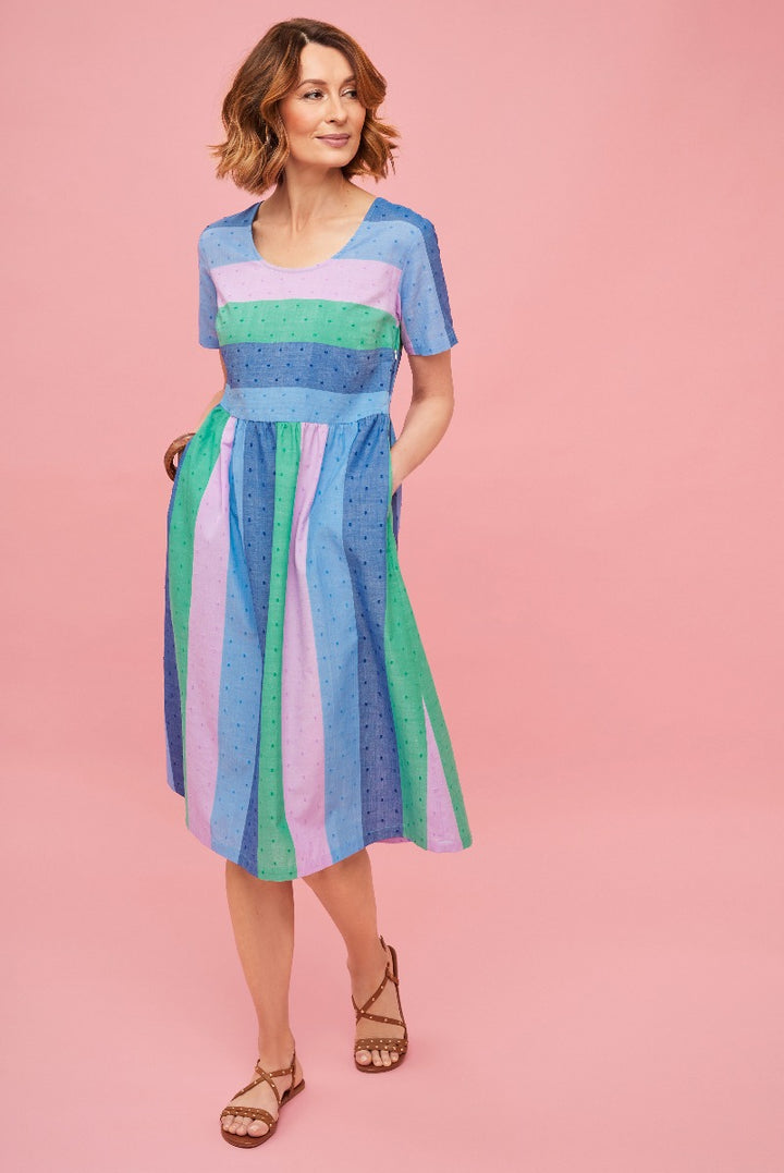 Lily Ella Collection colorful striped summer dress in shades of blue, green, and purple with polka dots, featuring a fitted waist and flared skirt, styled with brown strappy sandals on a pink background.