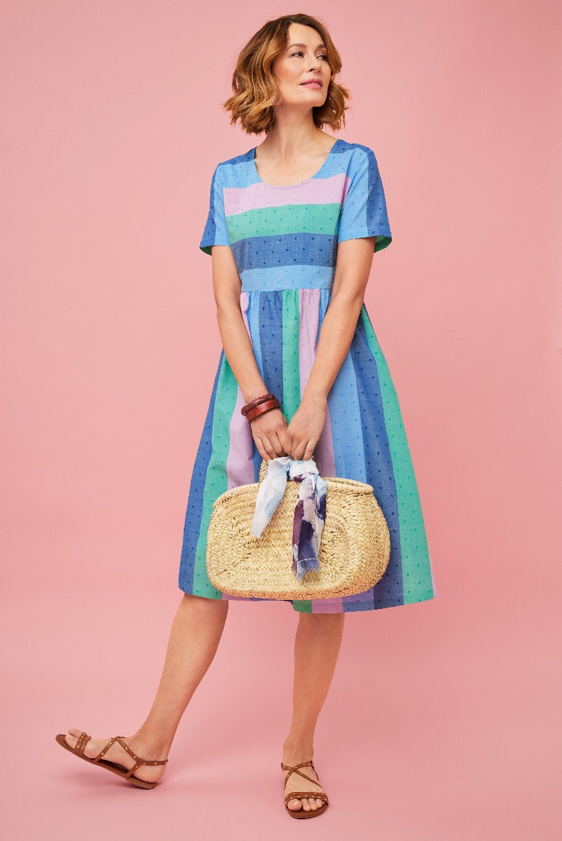 Lily Ella Collection summer dress, model wearing a blue and pastel striped knee-length dress with a scoop neckline, accessorized with a straw handbag and brown sandals, pink background.