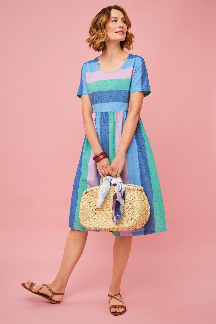 Lily Ella Collection summer dress, model wearing a blue and pastel striped knee-length dress with a scoop neckline, accessorized with a straw handbag and brown sandals, pink background.