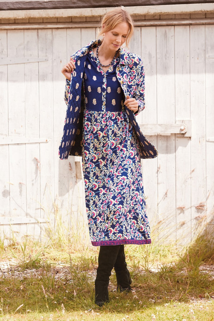 Woman modeling Lily Ella Collection navy floral dress with coordinating fringed jacket and black boots, outdoor setting with wooden background.