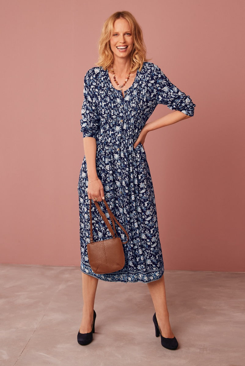 Lily Ella Collection navy blue floral print midi dress with three-quarter sleeves, paired with a brown leather shoulder bag and black heels, on a smiling model against a pink backdrop.