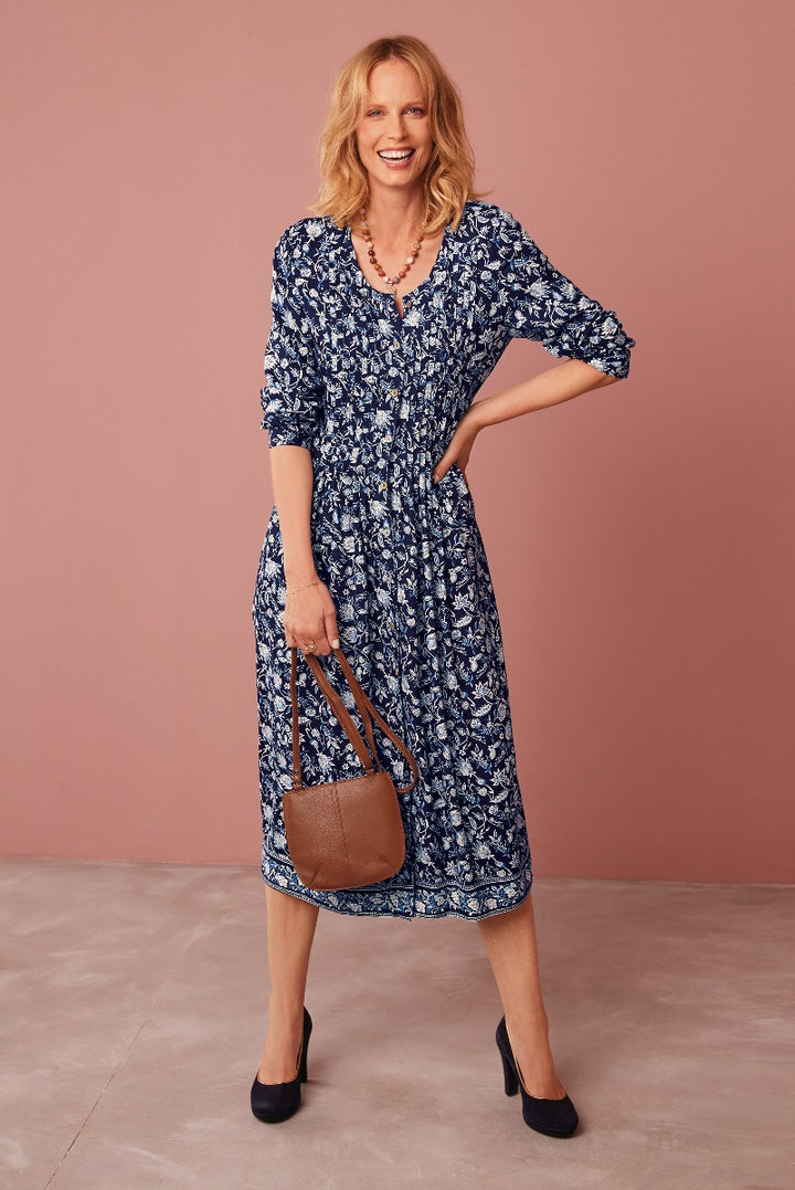 Lily Ella Collection navy blue floral print midi dress with three-quarter sleeves, paired with a brown leather shoulder bag and black heels, on a smiling model against a pink backdrop.