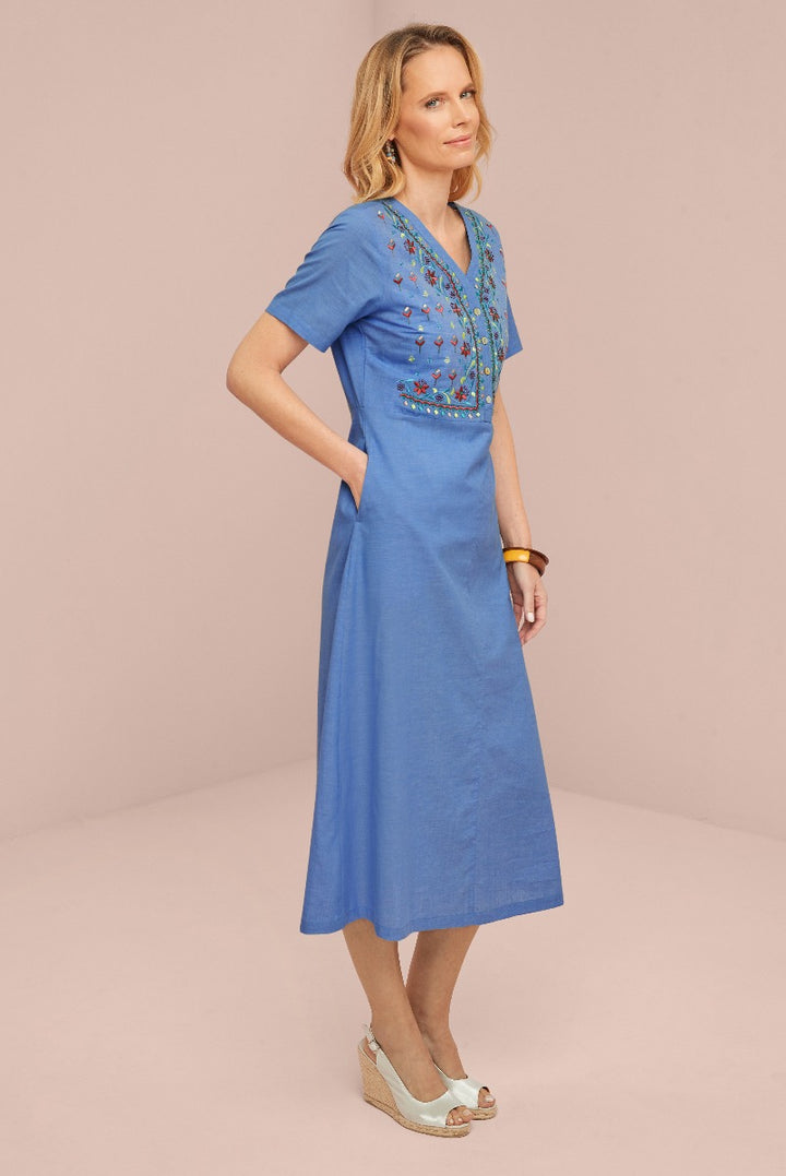 Lily Ella Collection blue mid-length dress with floral embroidery detailing, V-neck, short sleeves, and wedge sandals on model against a pink background.