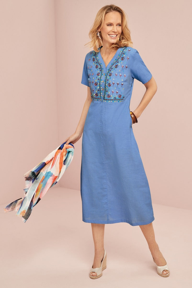 Lily Ella Collection blue mid-length dress with floral embroidery, V-neck, and short sleeves, accessorized with colorful scarf and beige wedge sandals.