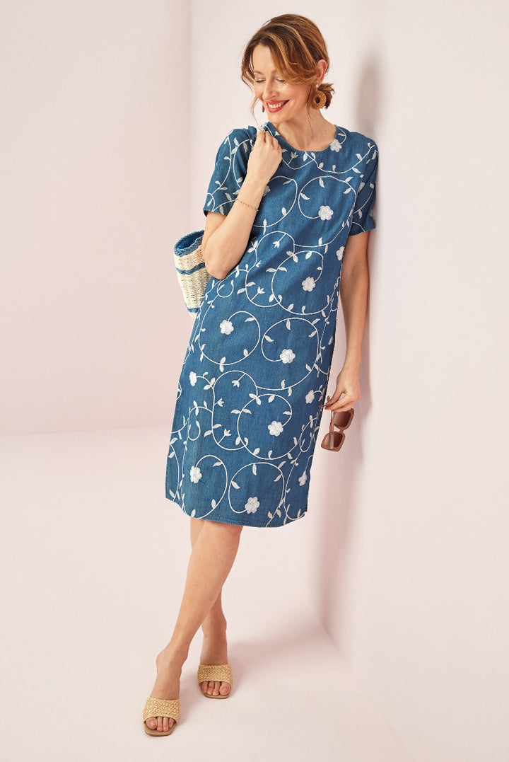 Lily Ella Collection blue floral patterned dress, stylish knee-length outfit with short sleeves and round neckline, accessorized with straw bag and heeled sandals, elegant summer fashion.