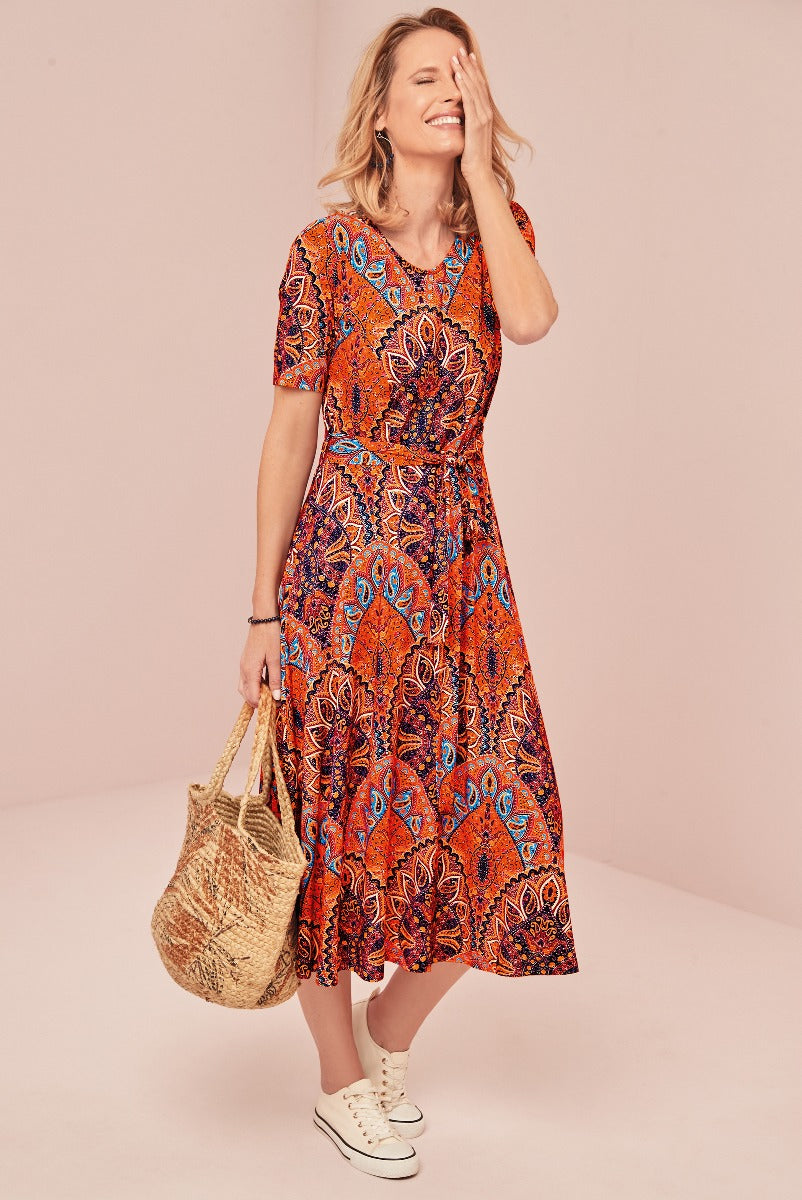 Lily Ella Collection vibrant orange and blue patterned midi dress with short sleeves, smiling woman accessorizing with a straw tote bag and white sneakers, casual summer outfit.