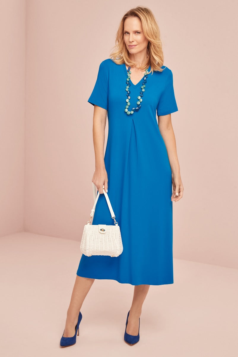 Lily Ella Collection elegant blue mid-length dress with V-neck, paired with white woven handbag and matching blue heels, accessorized with multi-tier green bead necklace.