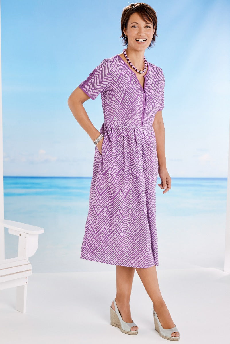 Lily Ella Collection summer fashion featuring cheerful woman in vibrant purple zig-zag pattern midi dress with v-neckline and short sleeves, accessorized with a necklace and bangle, posing against a serene beach backdrop.