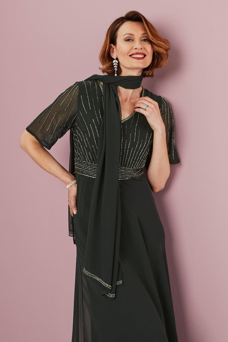 Lily Ella Collection elegant black evening dress with embellished accents, stylish sheer sleeves, and matching scarf accessory on a smiling model against a pink background