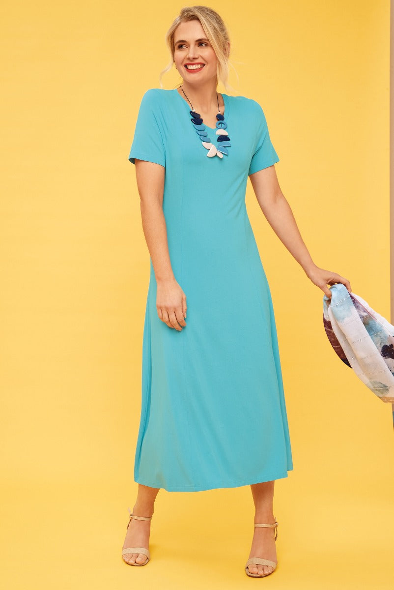 Lily Ella Collection turquoise midi dress with short sleeves and statement necklace, elegant casual wear, model holding pastel floral print scarf, stylish women's fashion against yellow background.
