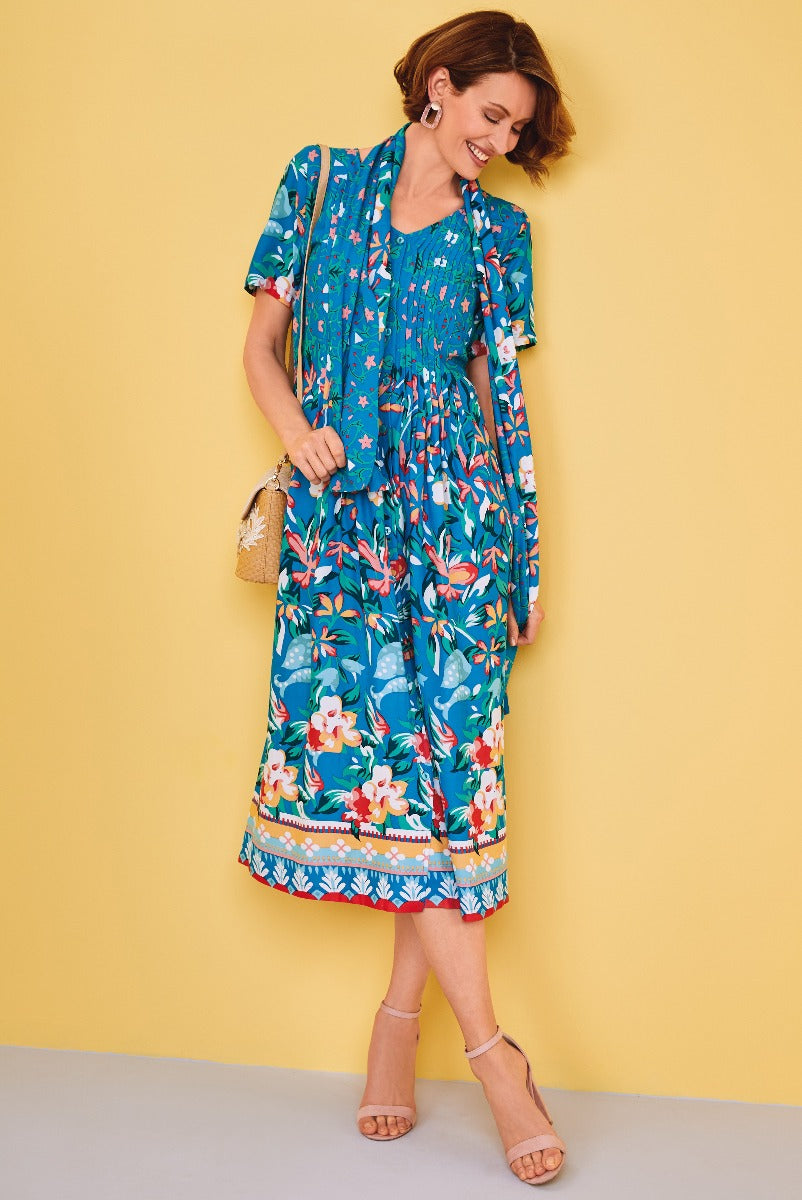 Lily Ella Collection floral print jumpsuit in blue with vibrant tropical pattern, stylish summer women's fashion, model showcasing sleeveless design with cinched waist and wide-leg trousers against yellow background.