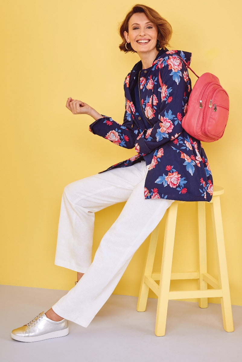 Lily Ella Collection floral print navy jacket with white trousers and coral pink backpack accessory on smiling model against yellow background