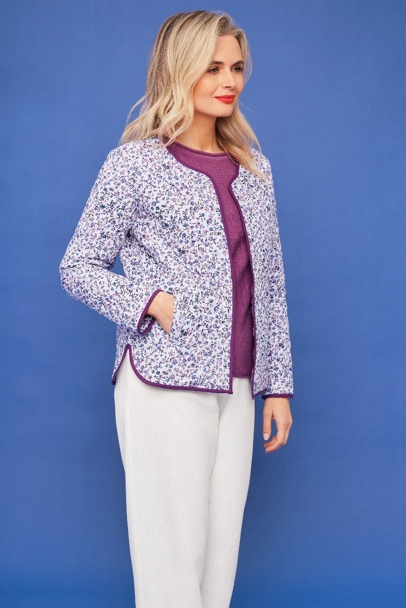 Lily Ella Collection stylish woman wearing a floral print purple jacket paired with a plum top and white trousers against a blue background