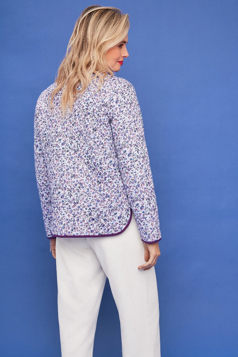 Lily Ella Collection floral print blouse in purple and white, elegant style, paired with white trousers, against a bright blue background.